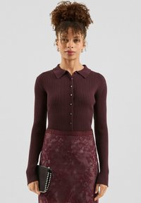 Burgunderfarbener gerippter Strickcardigan mit Polokragen, Knopfverschluss und langen Ärmeln, kombiniert mit einem Spitzenrock in Burgunder und einer schwarzen Clutch.