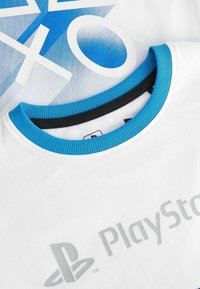 T-shirt blanc avec des accents bleus, comportant un design graphique des symboles et du logo PlayStation en gris. Matériau en coton avec col côtelé.