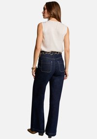 Jeans de mezclilla oscura de pierna ancha con costuras doradas, combinados con una blusa sin mangas clara. Cuenta con un cinturón circular dorado decorativo y dos bolsillos traseros.