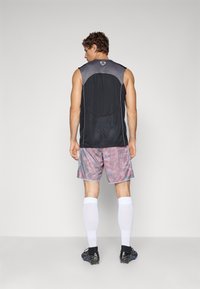 Mann som har på seg svart utenermet sports skjorte, grå og rød mønstrede shorts, hvite knelang sokker og svarte fotballsko, står med ryggen til.