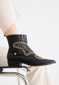 Botín negro de cuero con tachuelas doradas decorativas, punta afilada, cierre lateral con botón y un tacón bajo de bloque, combinado con pantalones beige deshilachados.
