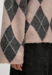 Hand rustend naast het lichaam, gekleed in een roze en grijs ruitpatroon fuzzy sweater met zwarte gestructureerde broek.