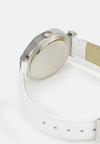 Silverfärgad metallklocka med vit läderrem. Har en strukturerad yta och en krona, visar en polerad finish med justerbara hål.