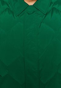 Giacca trapuntata verde con una texture lucida e liscia, ampio colletto e un motivo di cucitura diagonale. Chiusura frontale senza hardware visibile.