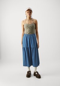 Haut ajusté vert olive avec des fines bretelles et des détails de couture, associé à une jupe en denim bleu à volants. Chaussures marron à bout fermé et chaussettes claires.