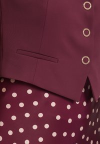 Burgunderfarbener Blazer mit einem goldenen runden Knopf, kombiniert mit einem burgunderfarbenen Rock mit hellrosa Polka-Dots. Glattes Material und maßgeschneidertes Design.