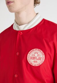 Red bomber jacket with a white, striped collar. Features a circular logo patch in white and red, and red snap buttons. Smooth texture.