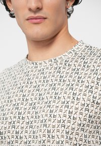 Primer plano de una persona que lleva una camisa con un patrón de letras "F" y "X" en negro y beige, repetidas sobre un fondo blanco.