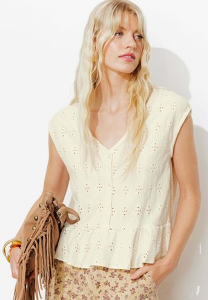 Femme blonde portant un chemisier sans manches écru en œillet et un pantalon fleuri, tenant un sac à main marron à franges, regardant sur le côté.