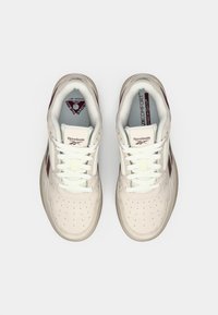Beigā krāsā Reebok sporta apavi ar baltām šņorēm, bordo akcentiem un mīkstajām zolēm, kuras marķētas ar "Lux Comfort" un "The Pump."