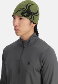 Gorro de punto verde con un diseño de araña negra, usado por una persona con una camiseta gris de cremallera corta con un logotipo de araña en el pecho.