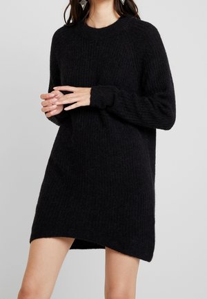 Kvinde iført en lang sort strikket sweaterkjole, hænder foldet, med geometriske sølvøreringe mod en ensfarvet lys baggrund.