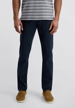 Mann trägt marineblaue, gerade geschnittene Hosen, ein grau-weiß gestreiftes Hemd und hellbraune Wildleder-Slipper, stehend vor weißem Hintergrund.