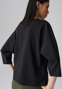 Schwarzes Oversized-Oberteil aus weichem Stoff mit ¾-Ärmeln, runder Ausschnitt und geradem Saum. Glatte Textur mit minimalen Details.
