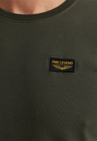 Tamnozeleni dukserica s okruglim izrezom s malim crnim i žutim logotipom PME Legend na prsima koji prikazuje krila i tekst.