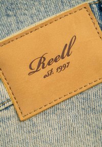 Ein brauner Lederpatch, der auf leicht blauer Jeans genäht ist, mit dem Markennamen "Reell" und "est. 1997" in Dunkelbraun eingeprägt.