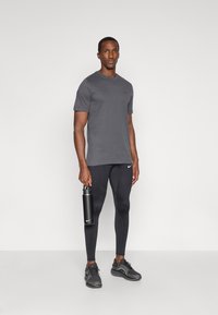 Γκρι αθλητικό μπλουζάκι με μικρό λογότυπο, συνδυασμένο με μαύρα κολάν με υφή και ένα μπουκάλι νερού της Nike στο χέρι.