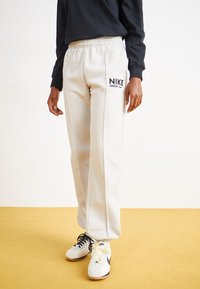 Világosszürke pamut melegítőnadrág rugalmas derékkal, fekete Nike logóval, fehér és fekete sportcipővel egy sárga padlón.