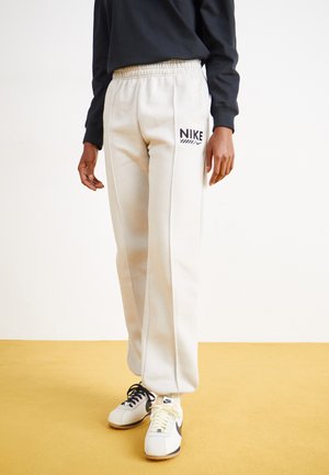 Pantaloni da tuta in cotone grigio chiaro con vita elasticizzata, caratterizzati dal logo Nike nero e abbinati a sneakers bianche e nere su un pavimento giallo.