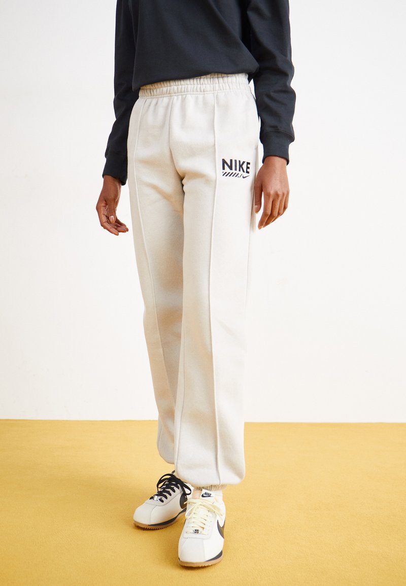 Világosszürke pamut melegítőnadrág rugalmas derékkal, fekete Nike logóval, fehér és fekete sportcipővel egy sárga padlón.