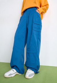 Blå cargobyxor gjorda av slitstarkt tyg, med flera fickor, en bred bendesign och elastiska muddar. I kombination med vita sneakers.