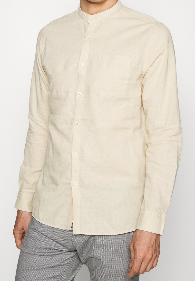 Homme portant une chemise légère beige à manches longues avec une poche poitrine et un pantalon gris à motifs, debout devant un fond uni.