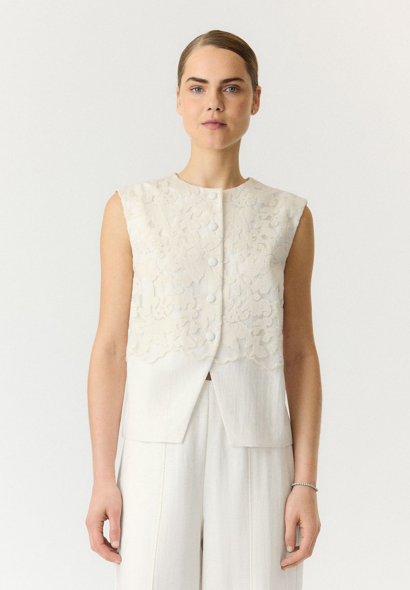 Femme à la peau claire et aux yeux bleus portant un gilet blanc sans manches texturé à motifs floraux et boutonné, ainsi qu’un pantalon blanc assorti, sur un fond uni.