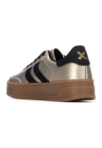 Zapatilla metálica dorada con rayas negras, parte superior texturizada y una suela de goma marrón. Presenta una lengüeta trasera negra con un acento dorado.