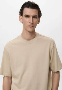 Tricou din bumbac bej, cu guler rotund și mâneci scurte, având o textură netedă și o croială lejeră. Fără modele sau accente vizibile.
