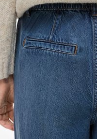 Plave traperice s stražnjim džepom, s teksturiranim završetkom i kontrastnim smešnim šavovima duž ruba džepa i pojasa.