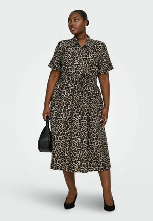 Donna che indossa un abito midi stampa leopardata con maniche corte, tiene una borsa nera e porta tacchi neri su uno sfondo semplice.