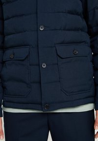 Marinblå puffjacka med vertikalt quiltat mönster, har två framfickor, svarta knappar och en lager-på-lager ljusgrön skjorta under.