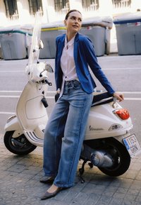 Balta Vespa motoroleris su skaidriu vėjo stiklu, pastatytas ant tekstūruotos plytelėmis grindinio dangos. Modelis vilki mėlyną švarką ir platėjančias džinsines kelnes, atsiremiantis į motorolerį.