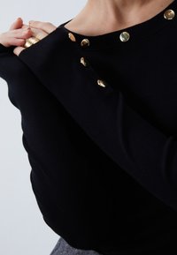 Haut noir à manches longues avec des boutons-pression dorés le long de l'encolure. Le tissu semble doux avec une texture côtelée. D étails comprenant des manches ajustées.