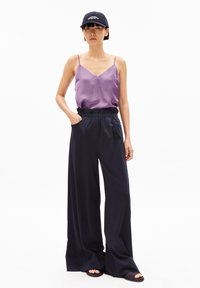 Lila Satin-Camisole mit dünnen Trägern, kombiniert mit navyblauen, weit geschnittenen Hosen mit einem elastischen Bund und seitlichen Taschen, abgerundet mit schwarzen Sandalen.