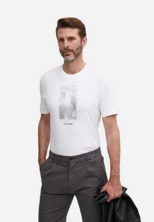 Camiseta blanca de algodón con un diseño gráfico y el texto "Calvin Klein", combinada con pantalones grises y sosteniendo una chaqueta de cuero negra.