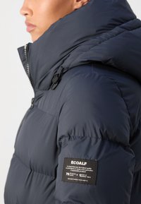 Chaqueta de plumas aislante de color azul marino con cuello alto, cierre de cremallera y etiqueta Ecoalf. Tejido texturizado y diseño elegante.