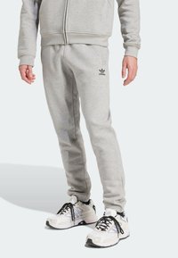 Šedé tepláky vyrobené z měkkého materiálu, s užším střihem, elastickým pasem a vyšívaným černým logem Adidas na levé noze.