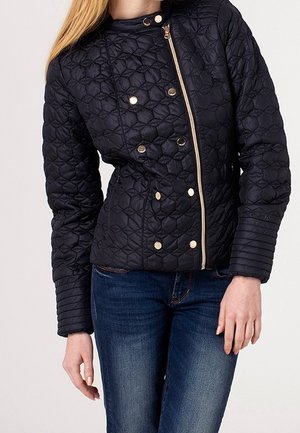 Kobieta ubrana w czarną pikowaną kurtkę z złotym zamkiem i guzikami, w połączeniu z niebieskimi dżinsami, stoi na jasnym tle.