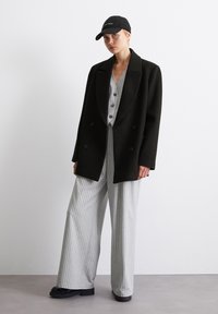 Černý oversized dvouřadový kabát přes šedý pruhovaný široký overal, doplněný černou čepicí a robustními čern ými platformovými botami.