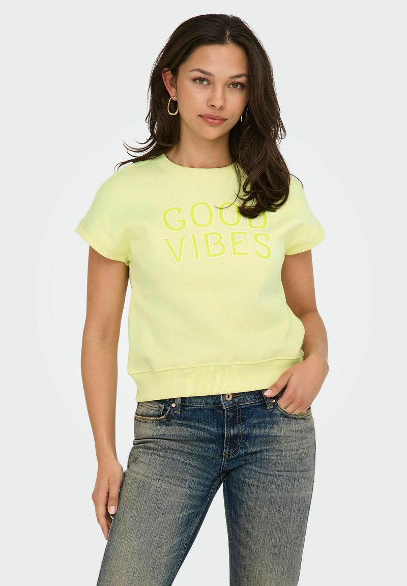 Jeune femme aux longs cheveux noirs portant des boucles d'oreilles en or en forme de créoles, un t-shirt jaune à manches courtes avec l'inscription « Good Vibes » et un jean bleu, debout avec une main dans la poche.