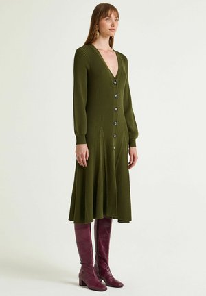 Knee-length olive green ribbed dress with a V-neck, long sleeves, and front buttons. Worn with knee-high burgundy textured boots.