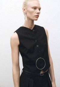 Top senza maniche nero con bottoni e design aderente, abbinato a una sottile cintura nera. Collana con pendente circolare e orecchini a goccia in materiale trasparente.