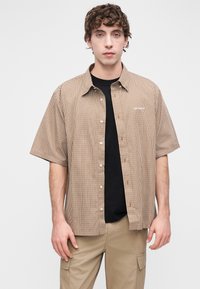 Κοντομάνικο πουκάμισο με ρίγες σε καφέ και κρεμ χρώμα, με κουμπιά μπροστά, από κάτω μαύρο T-shirt. Φοράται με ανοιχτό μπεζ παντελόνι.