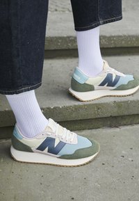 Sneakers med en blandning av ljusblå, grön och gräddfärgad material, med en framträdande mörkblå "N"-logotyp och en strukturerad vit sula.