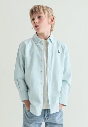 Blond jongen met lichtblauw gestreept overhemd over een wit T-shirt en een blauwe spijkerbroek, staand met handen in de zakken tegen een effen achtergrond.