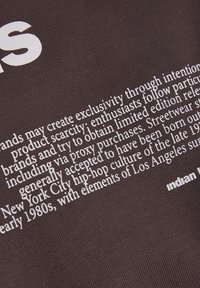 Brunt tyg med vit text, med en typografisk design. Texten innehåller information om produktbrist och streetwear-kultur.