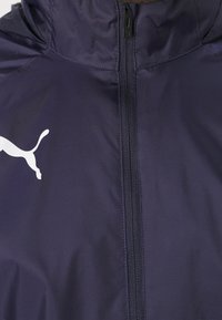 Puma-takki tummansinisestä kankaasta, jossa on täyspitkä vetoketju edessä ja valkoinen logo vasemmalla puolella. Sileä pinta ja kevyt muotoilu.
