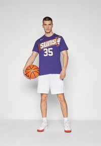 Paarse basketbaltruit met het "Suns" logo en nummer 35, gecombineerd met witte shorts en grijs-oranje sneakers. Houdt een basketbal vast.