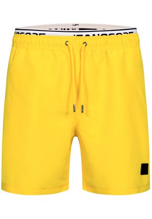 Muskularny mężczyzna boso, uśmiechnięty i patrzący w dół, ubrany w jasne żółte kąpielówki ze czarnym sznurkiem na jednolitym jasnym tle.